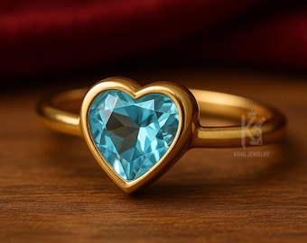 May include: A gold ring featuring a heart-shaped, light blue gemstone. The gemstone is set in a gold bezel, and the ring band is also gold. The ring is displayed on a wooden surface. The text "KING JEWELRY" is visible.