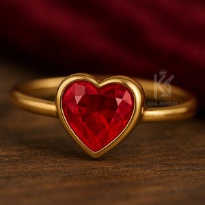 May include: A gold ring featuring a heart-shaped, faceted, red gemstone. The gemstone is set in a gold bezel. The ring is displayed on a wooden surface. The words "KING JEWELRY" are visible.