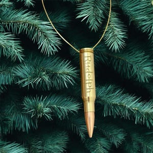 May include: A gold-colored bullet pendant with the name "Rudolph" engraved vertically. The pendant hangs from a thin gold chain against a backdrop of green pine needles.