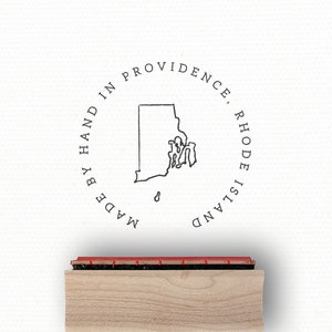 May include: Wooden stamp with a brown handle. The stamp features a black outline of the state of Rhode Island with the text "Made by Hand in Providence, Rhode Island".
