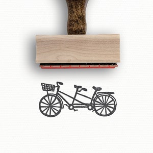 May include: A wooden rubber stamp with a brown handle and a rectangular base. The stamp features a black and white illustration of a tandem bicycle with a basket on the front.