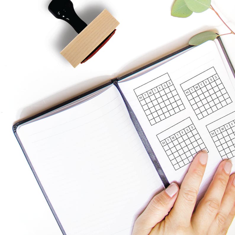 Puede incluir: Un cuaderno blanco con cuatro cuadr&iacute;culas de calendario semanal en blanco impresas en las p&aacute;ginas. Las cuadr&iacute;culas est&aacute;n etiquetadas con los d&iacute;as de la semana: L, Ma, Mi, J, V, S, D. Un sello de madera con un mango negro se muestra encima del cuaderno.