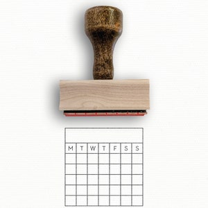 Puede incluir: Un sello de madera con una almohadilla de tinta roja y un dise&ntilde;o de cuadr&iacute;cula de calendario. La cuadr&iacute;cula del calendario tiene los d&iacute;as de la semana etiquetados como M, T, W, T, F, S, S.