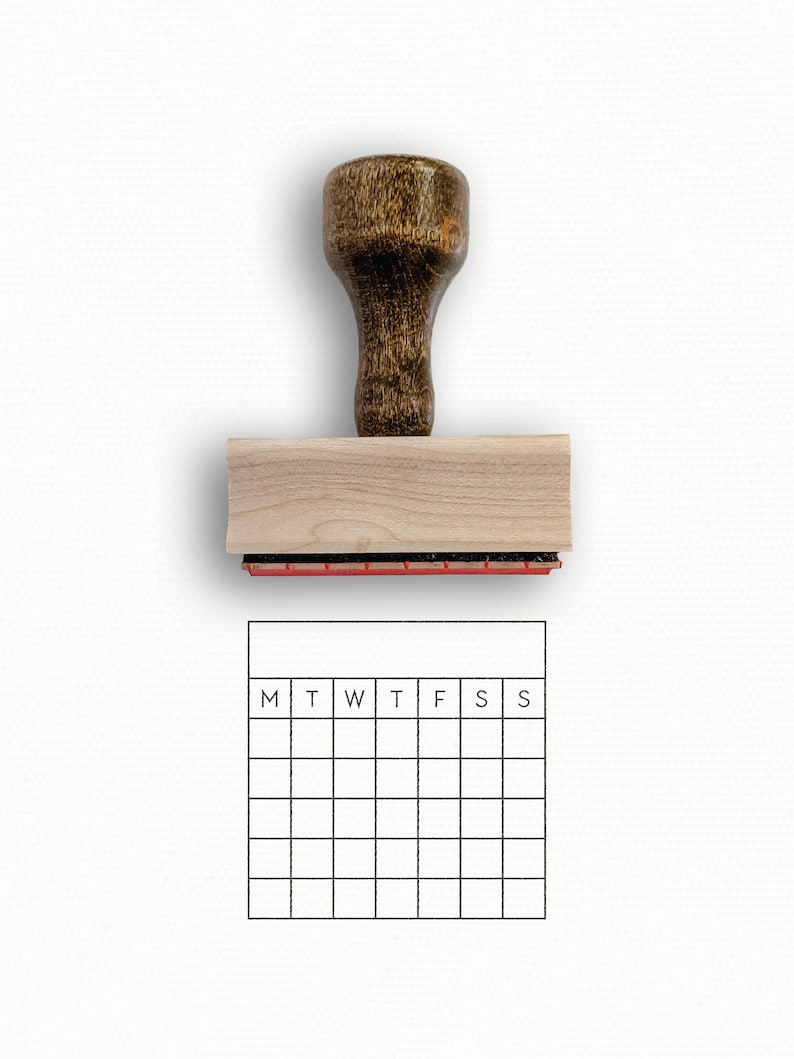 Puede incluir: Un sello de madera con una almohadilla de tinta roja y un dise&ntilde;o de cuadr&iacute;cula de calendario. La cuadr&iacute;cula del calendario tiene los d&iacute;as de la semana etiquetados como M, T, W, T, F, S, S.