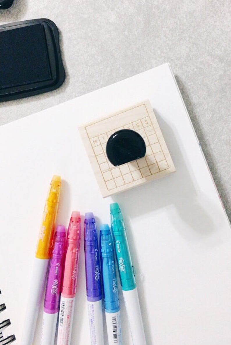 Puede incluir: Un sello de madera con un patr&oacute;n de cuadr&iacute;cula y las letras "MT" y "SS" impresas en &eacute;l. El sello est&aacute; sentado sobre una superficie blanca junto a seis rotuladores de colores. Los rotuladores son todos blancos con un barril de pl&aacute;stico transparente y una punta de color. Los colores de los rotuladores son amarillo, rosa, morado, azul y turquesa.