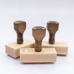 May include: Three wooden stamps with brown handles and rectangular bases. The bases are engraved with the text "Bakery, 7001 E. 1st Ave, Tacoma, WA 98410".