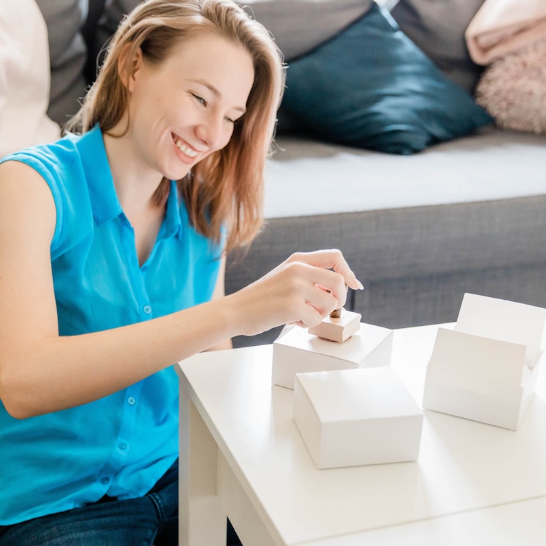 May include: A woman in a blue shirt is packaging a small white box with a brown lid. The box is sitting on a white table. There are two other white boxes on the table.