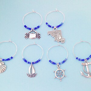 May include: Set of six blue and silver wine glass charms with nautical themed charms, including a crab, a sailboat, a seashell, a ship's wheel, a state of Maryland outline, and an anchor.