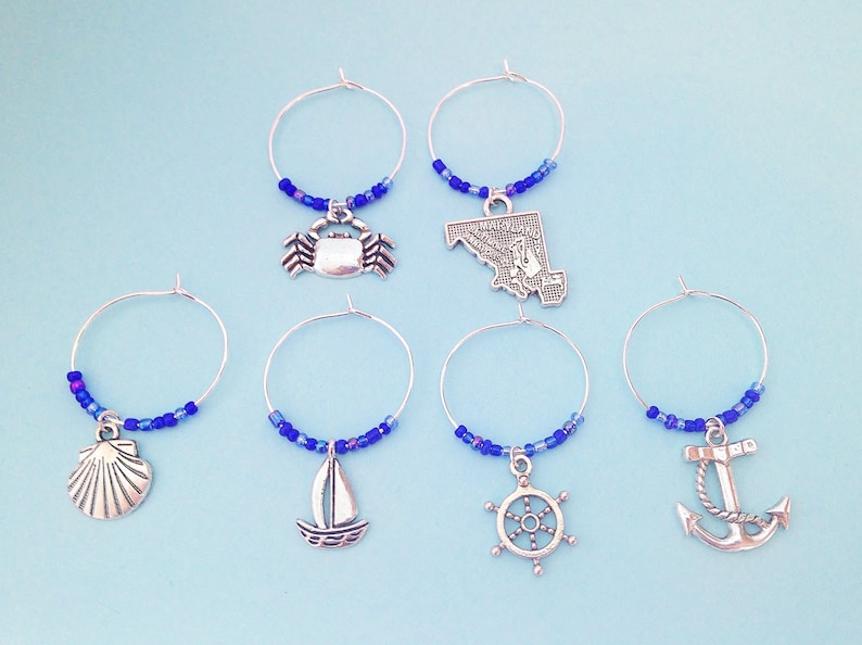 May include: Set of six blue and silver wine glass charms with nautical themed charms, including a crab, a sailboat, a seashell, a ship's wheel, a state of Maryland outline, and an anchor.