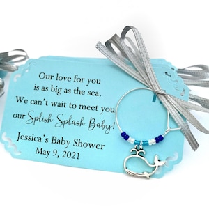 May include: A light blue baby shower favor tag with a silver whale charm and blue and white beads. The tag reads "Our love for you is as big as the sea. We can't wait to meet you our Splish Splash Baby! Jessica's Baby Shower May 9, 2021"