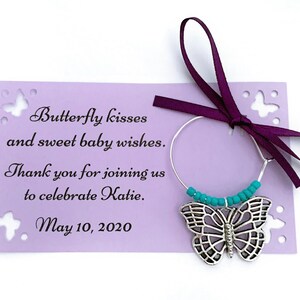 May include: A purple card with a butterfly design and the text "Butterfly kisses and sweet baby wishes. Thank you for joining us to celebrate Katie. May 10, 2020". A silver butterfly charm with turquoise beads hangs from a silver wire.