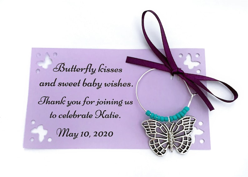 May include: A purple card with a butterfly design and the text "Butterfly kisses and sweet baby wishes. Thank you for joining us to celebrate Katie. May 10, 2020". A silver butterfly charm with turquoise beads hangs from a silver wire.