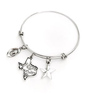 May include: Silver-toned bangle bracelet with three charms: a cowboy hat, a Texas state outline, and a five-pointed star. The Texas charm features the state's outline with the word "Texas" and city names. The bracelet is on a white background.