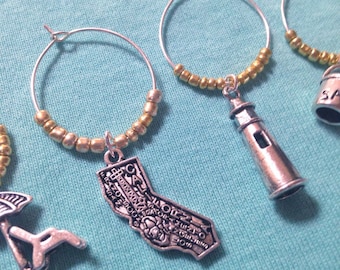May include: Four silver wine glass charms with gold beads. Each charm features a different design: a beach umbrella, a lighthouse, an outline of California, and a bucket with the word "Sand" on it.