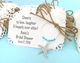 Może przedstawiać: Biała kartka z koronkową obwódką i brązową wstążką ze sznurka. Na kartce widnieje tekst "Cheers to love, laughter & happily ever after! Katie's Bridal Shower June 2, 2019". Srebrny zawieszka w kształcie rozgwiazdy zwisa z niebieskiego, koralikowego charmsa do kieliszka do wina.