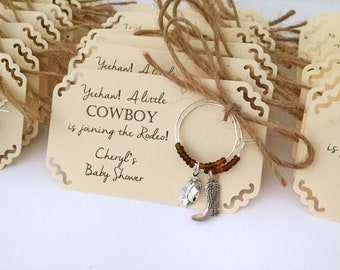 Op de afbeelding: Een set van 10 beige papieren wijnbekercharms met een bruine touw strik. Elke charme heeft een zilveren cowboylaars en hoed charme, en de tekst "Yeehaw! A little COWBOY is joining the Rodeo! Cheryl's Baby Shower".