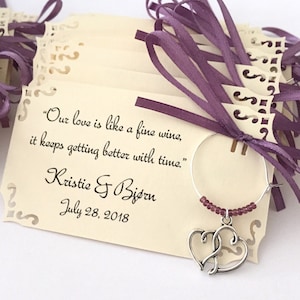 May include: A set of ivory wedding favour tags with a purple ribbon and a silver charm. The tags feature the quote "Our love is like a fine wine, it keeps getting better with time." and the names "Kristie & Bjørn" with the date "July 28, 2018".