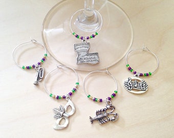 May include: Set of four silver wine glass charms with purple and green beads. Each charm features a different New Orleans themed design: a trumpet, a musical note, a mask, and a lobster.