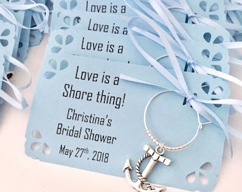 Może przedstawiać: Jasnoniebieska etykieta na prezent na wieczór panieński z tekstem "Love is a Shore thing!" i "Christina's Bridal Shower May 27th, 2018". Srebrny zawieszka w kształcie kotwicy jest przymocowana do srebrnego zawieszki do kieliszka do wina.