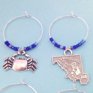 May include: Two silver wine glass charms with blue beads. One charm features a silver crab and the other charm features a silver outline of the state of Maryland with the text "MARYLAND" and the cities "BALTIMORE" and "ANNAPOLIS".