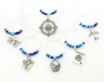 May include: Set of six blue and silver wine glass charms with travel themed charms. The charms include a compass, airplane, camera, suitcase, globe, and passport.