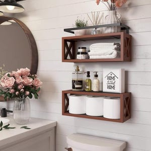 May include: A bathroom scene with three wooden shelves mounted on a white wall. The shelves hold white towels, toiletries, and toilet paper rolls. A sign reads "Home Sweet Home". A round mirror and a vase of pink roses are also visible.