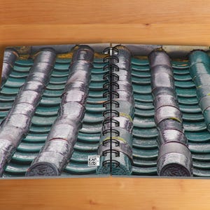 May include: A spiral-bound notebook open to a detailed image of a traditional Asian-style roof. The roof tiles are a teal green color, with cylindrical, silver-colored accents. The notebook is on a wooden surface.