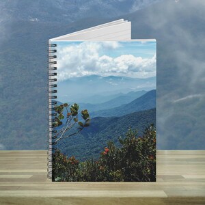 Puede incluir: Un cuaderno con espiral con un paisaje de montaña en la portada. La imagen presenta capas de montañas azules bajo un cielo nublado, con follaje verde en primer plano. El cuaderno está abierto, revelando páginas blancas.