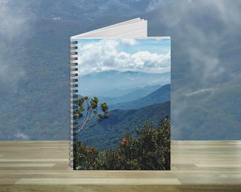 May include: A spiral-bound notebook with a scenic mountain landscape on the cover. The image features layers of blue mountains under a cloudy sky, with green foliage in the foreground. The notebook is open, revealing white pages.