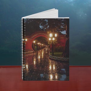 Könnte beinhalten: Spiralgebundenes Notizbuch mit einem Cover, das eine Nachtszene eines roten Torbogens und eines mit Straßenlaternen beleuchteten Weges zeigt. Das Bild zeigt eine nasse, reflektierende Oberfläche, wobei die Lichter einen warmen Schein vor dem dunklen Hintergrund erzeugen.