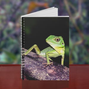 May include: Spiral-bound notebook featuring a vibrant image of a green lizard perched on a branch. The lizard's head and front legs are visible, set against a dark background. The notebook's pages are white, and the cover is glossy.
