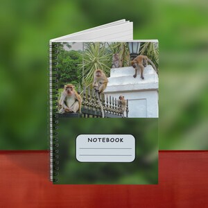 May include: Spiral-bound notebook with a green cover featuring a photograph of monkeys. The notebook is open, revealing lined pages. The cover has the word "NOTEBOOK" in a white rectangle.