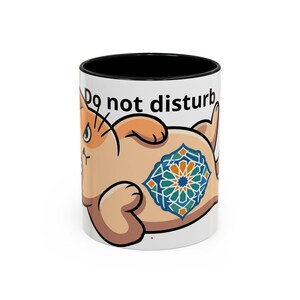 Puede incluir: Taza de cerámica blanca con interior negro, con un diseño de gato de dibujos animados. El gato es de color marrón claro con un patrón geométrico azul, verde y naranja en la espalda. Las palabras "Do not disturb" están impresas en la taza.