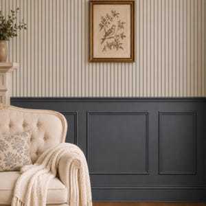 Op de afbeelding: Een cr&egrave;mekleurige gecapitonneerde bank met een cr&egrave;mekleurige kabelgebreide plaid en een kussen met patroon. De muur heeft beige en blauw gestreept behang boven donkergrijze lambrisering, met een ingelijste vogelprent in het midden.