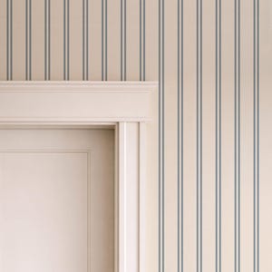 Op de afbeelding: Behang met een patroon van dunne, verticale, donkerblauwe strepen op een effen cr&egrave;mekleurige achtergrond. Het behang is aangebracht op een muur rondom een wit deurkozijn met een decoratieve lijst aan de bovenzijde.