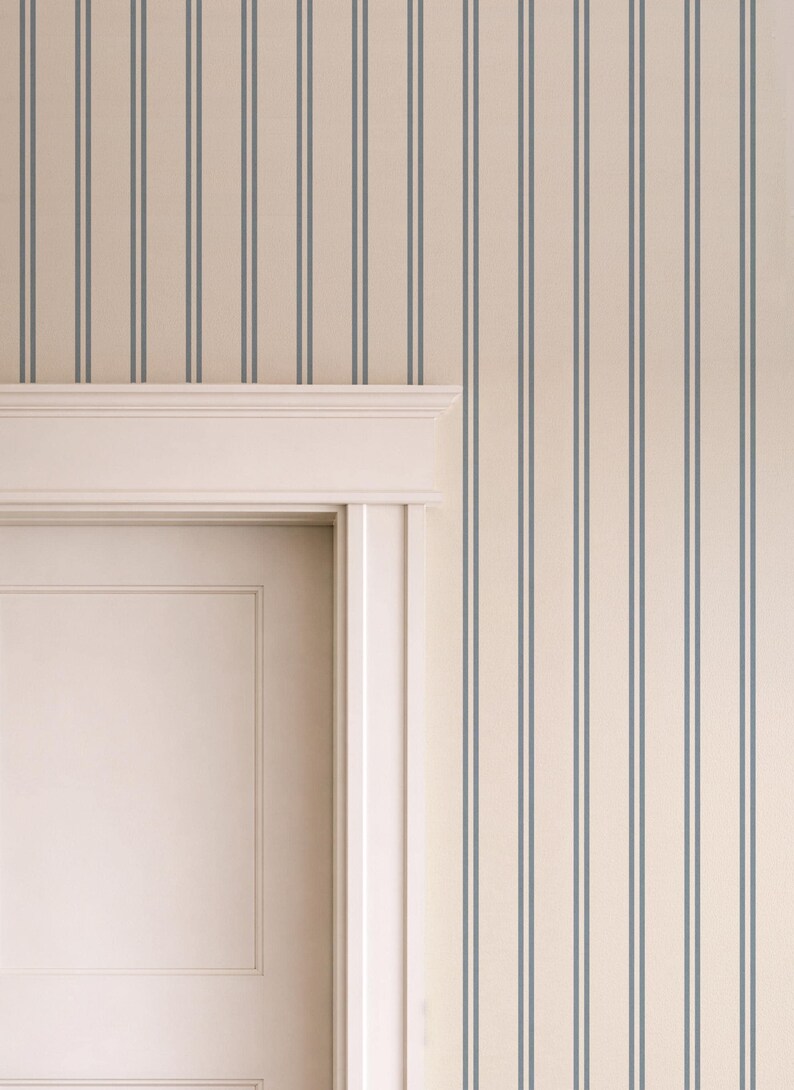 Op de afbeelding: Behang met een patroon van dunne, verticale, donkerblauwe strepen op een effen cr&egrave;mekleurige achtergrond. Het behang is aangebracht op een muur rondom een wit deurkozijn met een decoratieve lijst aan de bovenzijde.