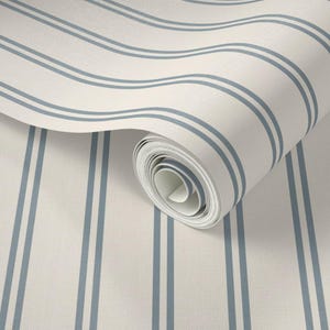 Op de afbeelding: Een rol behang met een patroon van verticale, parallelle blauwe strepen op een effen gebroken witte achtergrond. Het behang is gedeeltelijk uitgerold, waardoor de binnenste lagen van de rol en het consistente gestreepte ontwerp zichtbaar zijn.