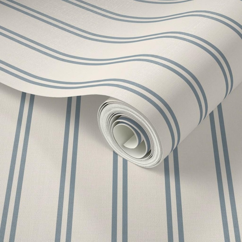 Op de afbeelding: Een rol behang met een patroon van verticale, parallelle blauwe strepen op een effen gebroken witte achtergrond. Het behang is gedeeltelijk uitgerold, waardoor de binnenste lagen van de rol en het consistente gestreepte ontwerp zichtbaar zijn.