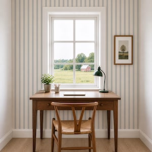 Op de afbeelding: Een houten bureau met drie lades en een bijpassende houten stoel met een gevlochten zitting. Op het bureau staan een kleine plant in een pot, een pennenhouder en een groene bureaulamp. De muur is voorzien van behang met blauw-witte verticale strepen.