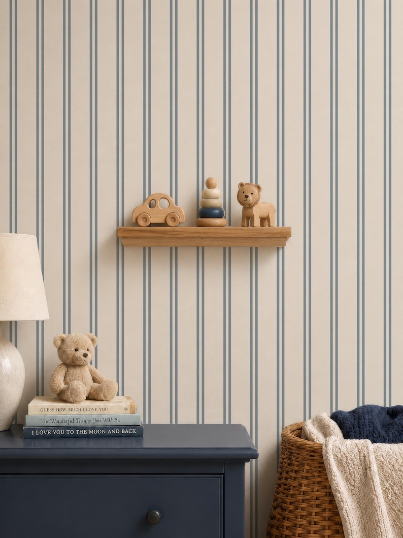 Op de afbeelding: Een muur met beige en blauw verticaal gestreept behang, een houten plank met een houten auto, een stapelspeeltje en een leeuwenfiguur. Daaronder staat een donkerblauwe ladekast met een teddybeer en boeken Guess How Much I Love You.