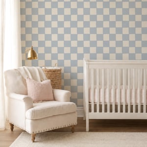 May include: A nursery room featuring a wall with a light blue and cream checkered pattern, a white crib, a cream-colored upholstered armchair with a light pink throw pillow, a brass lamp, a woven basket, and a textured cream area rug.