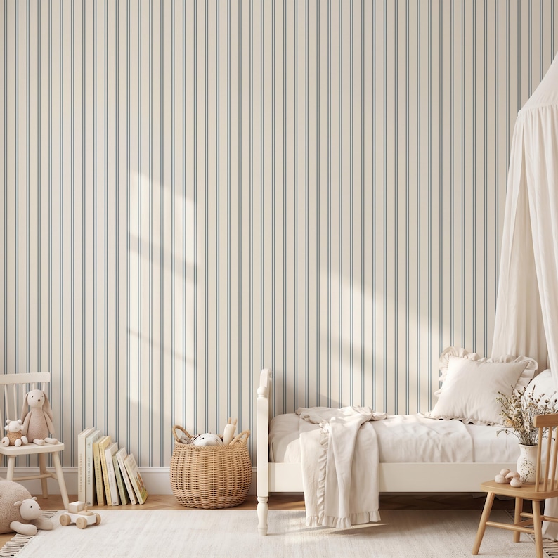 Op de afbeelding: Behang met een herhalend patroon van dunne, verticale blauwe strepen op een cr&egrave;mekleurige achtergrond. Het ontwerp bedekt een muur achter een bed met wit beddengoed, een rieten mand, een houten stoel en een verzameling boeken.