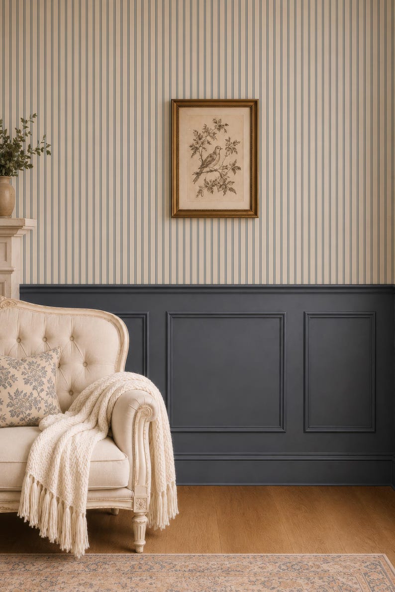 Op de afbeelding: Een cr&egrave;mekleurige gecapitonneerde bank met een cr&egrave;mekleurige kabelgebreide plaid en een kussen met patroon. De muur heeft beige en blauw gestreept behang boven donkergrijze lambrisering, met een ingelijste vogelprent in het midden.