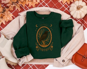 May include: Dark green sweatshirt featuring a gold-colored oval design with a raven perched on a tree branch. The design includes crescent moons and stars. The sweatshirt is displayed on a beige jacket and a red plaid surface.