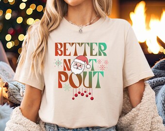 May include: Beige t-shirt with the text "BETTER NOT POUT" in red, green, and white. The design includes a Santa Claus face and snowflakes. Red ornaments hang below the text. A festive holiday shirt.