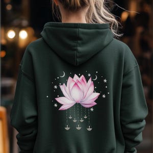Puede incluir: Sudadera con capucha verde oscuro con un diseño de flor de loto rosa y blanco en la espalda. El diseño incluye estrellas, lunas y formas colgantes de velas. La sudadera tiene capucha y un ajuste relajado.