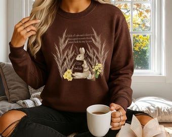 May include: A brown sweatshirt featuring a graphic of a rabbit holding a sign that says "Stay awhile." The design includes floral and botanical elements, with text that reads "In fields of calm and colors true..."