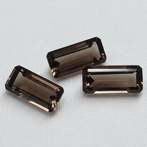 May include: Three rectangular, faceted gemstones with a translucent, dark brown hue. Each stone features a step-cut design with sharp, clean edges and a polished surface that reflects light, showcasing a consistent deep smoky colour throughout.