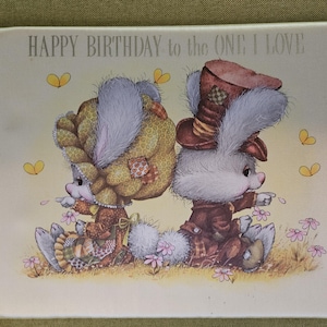Op de afbeelding: Een vintage verjaardagskaart met de tekst "HAPPY BIRTHDAY to the ONE I LOVE". De kaart toont twee cartoonkonijnen gekleed in Victoriaanse kleding, omringd door bloemen en vlinders. De konijnen zitten rug aan rug.