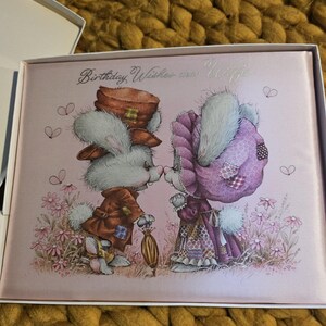 Puede incluir: Una tarjeta de felicitación rosa con una ilustración de dibujos animados de dos conejos besándose. Los conejos visten ropa vintage y están rodeados de flores. El texto "Birthday Wishes dear Wife" está escrito en la tarjeta.